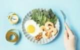 Female hand holds a fork next to a plate of food and centimeter tape, concept of a keto diet.