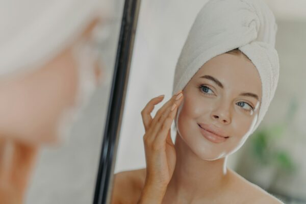 Beautiful woman touches face gently, looks at her reflection in mirror, enjoys softness of skin