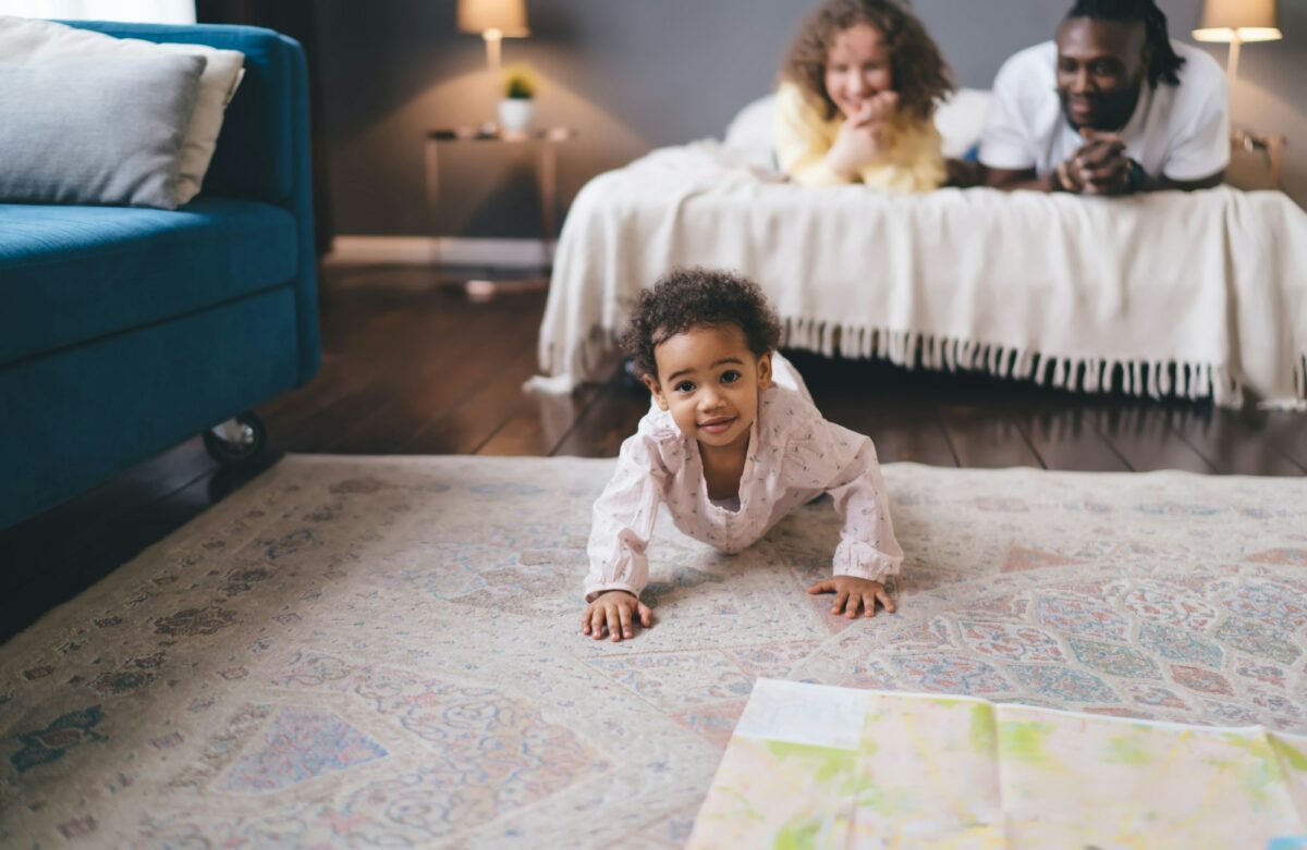 Cute black girl crawling on floor against background of parent lying on bed