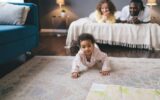 Cute black girl crawling on floor against background of parent lying on bed