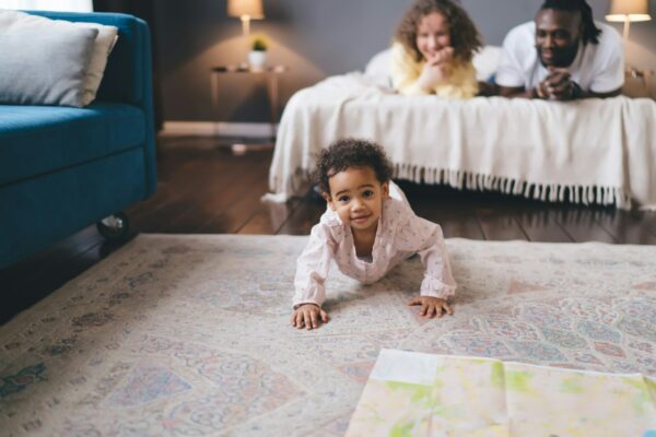Cute black girl crawling on floor against background of parent lying on bed