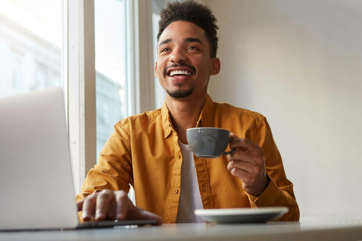 Photo of laughting African American attracrive guy, sits in a cafe, works at a laptop and drinks