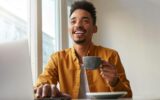 Photo of laughting African American attracrive guy, sits in a cafe, works at a laptop and drinks
