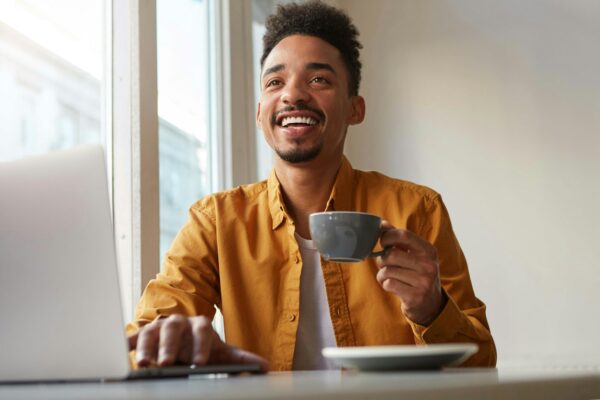 Photo of laughting African American attracrive guy, sits in a cafe, works at a laptop and drinks