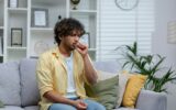 Sick cold man sitting alone on sofa at home, hispanic has bronchitis coughs shortness of breath