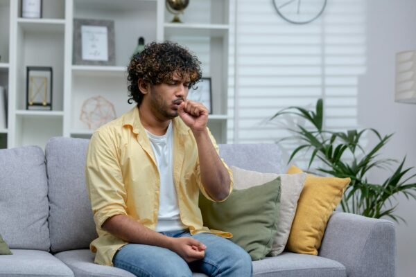 Sick cold man sitting alone on sofa at home, hispanic has bronchitis coughs shortness of breath